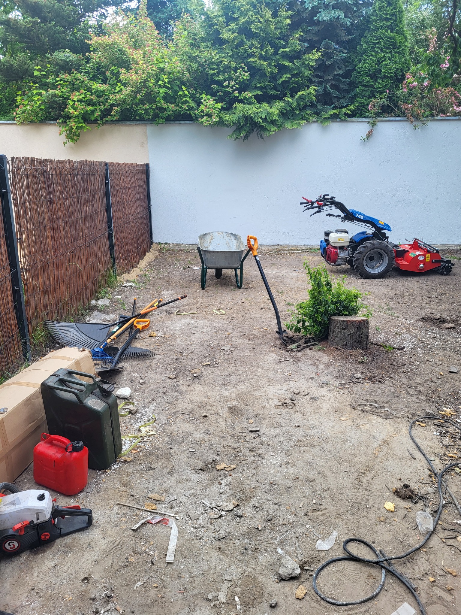 Teren po wycince drzewa z widocznym pniem, kosiarką rotacyjną, taczką, kanistrami, pilarką, grabiami i łopatą, otoczony płotem z wikliny i białym murem.