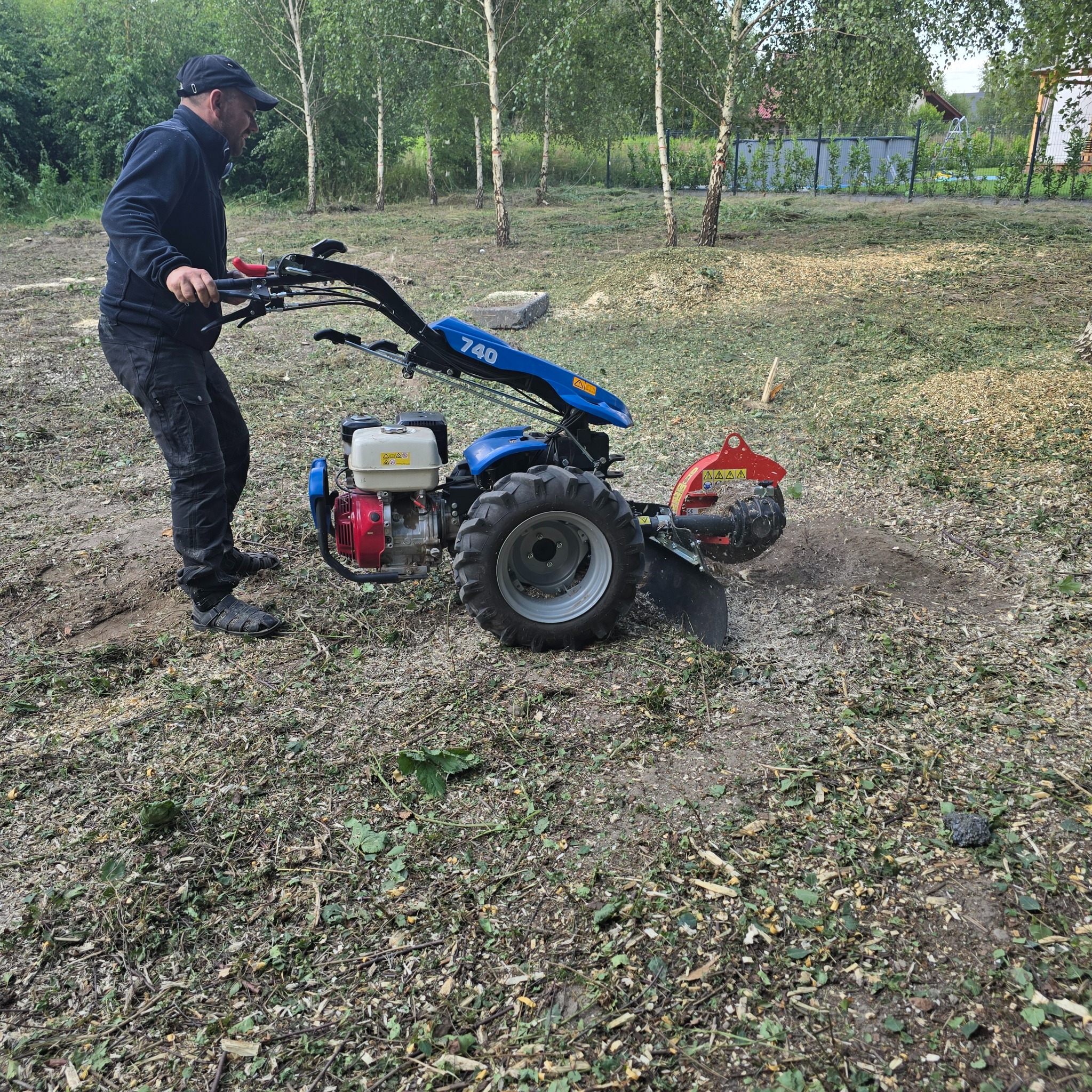 Mężczyzna obsługuje frezarkę do pni drzew na działce z widocznymi śladami po wycince. W tle drzewa i zabudowania.