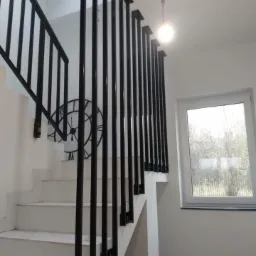 Białe schody z czarną metalową balustradą o minimalistycznym designie, z widocznym zegarem na ścianie i oknem w tle.