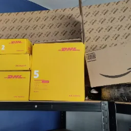 Stos żółtych kartonów DHL Express o różnych rozmiarach i brązowe pudełko Amazon na metalowym regale, gotowe do wysyłki.