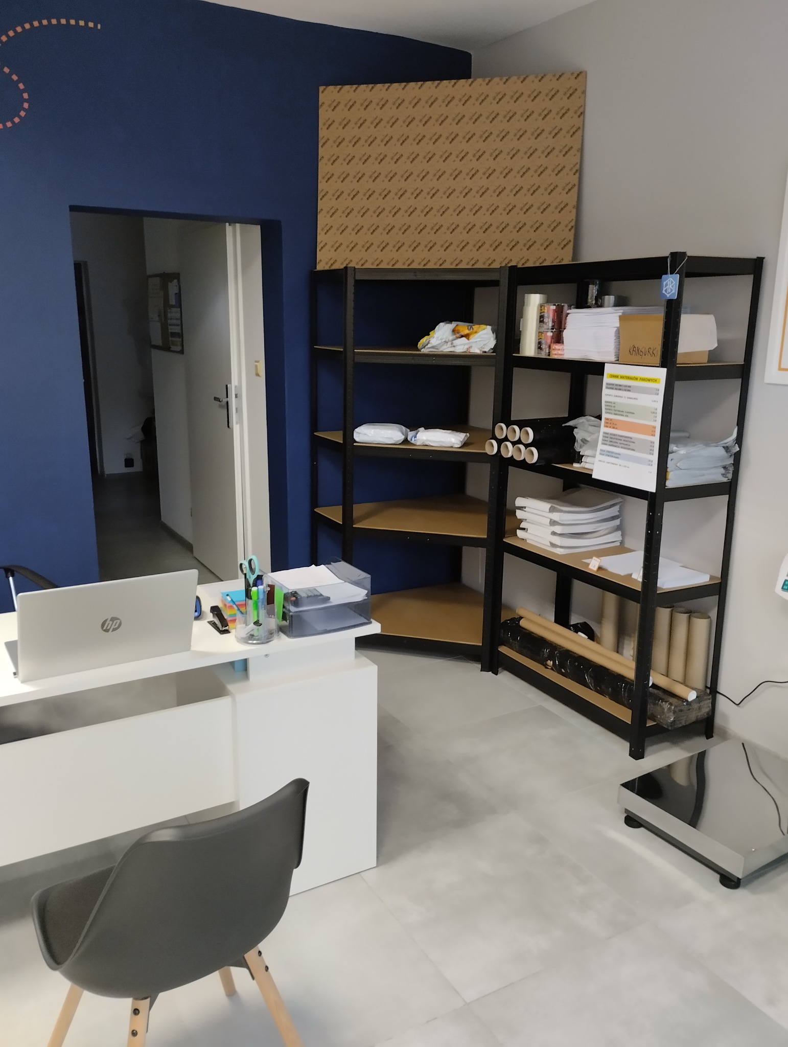 Biuro firmy transportowej: biurko z laptopem, regał z materiałami do pakowania, waga do paczek.