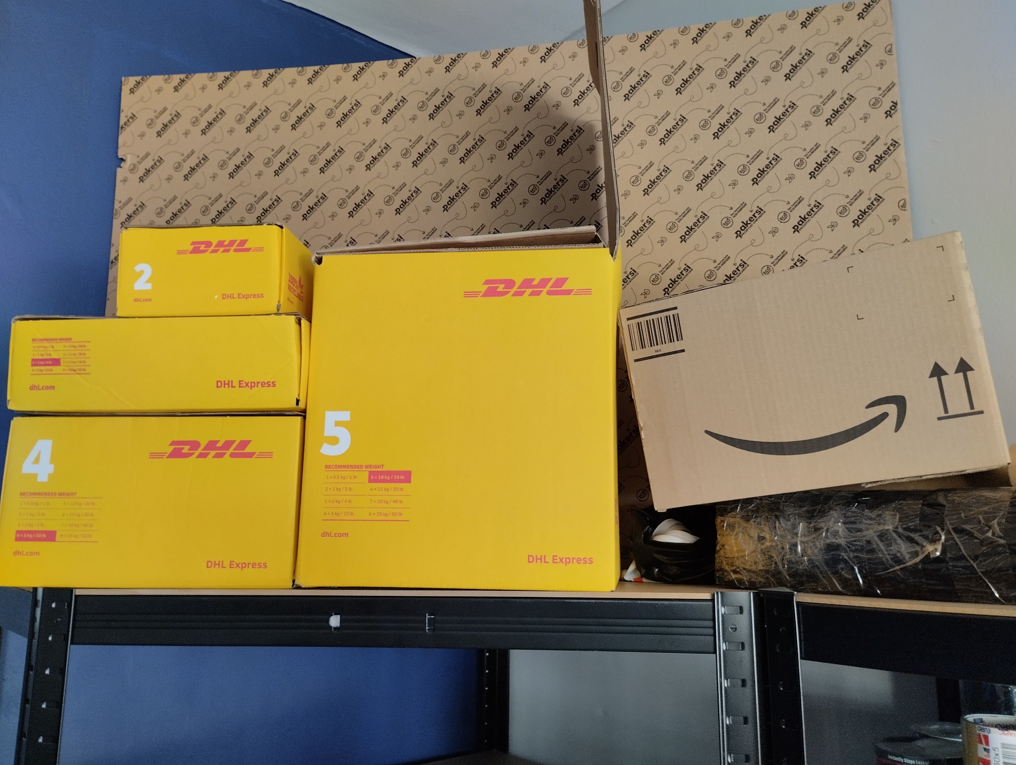 Stos żółtych kartonów DHL Express o różnych rozmiarach i brązowe pudełko Amazon na metalowym regale, gotowe do wysyłki.