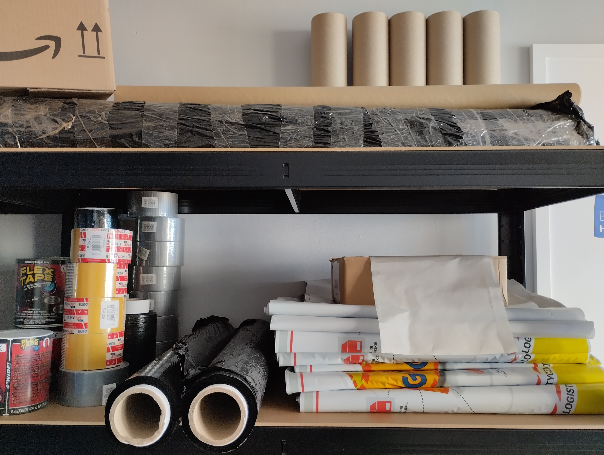 Półka magazynowa z materiałami do pakowania: rolki folii stretch, taśmy klejące, kartony i tuby tekturowe.
