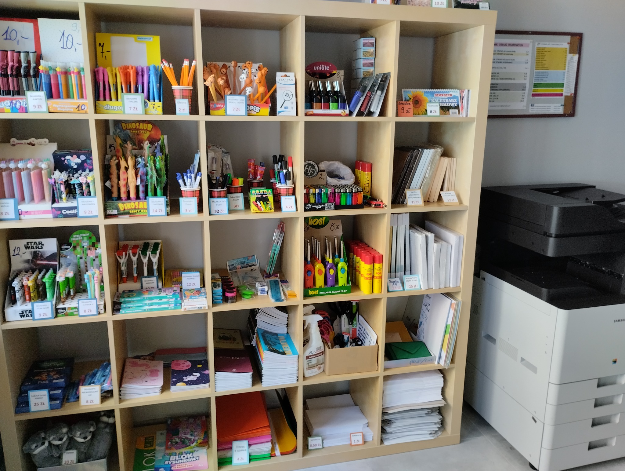 Regał z artykułami biurowymi i szkolnymi, w tym długopisami, ołówkami, markerami, zeszytami, kalendarzami i innymi akcesoriami. Obok regału stoi duża biała drukarka.