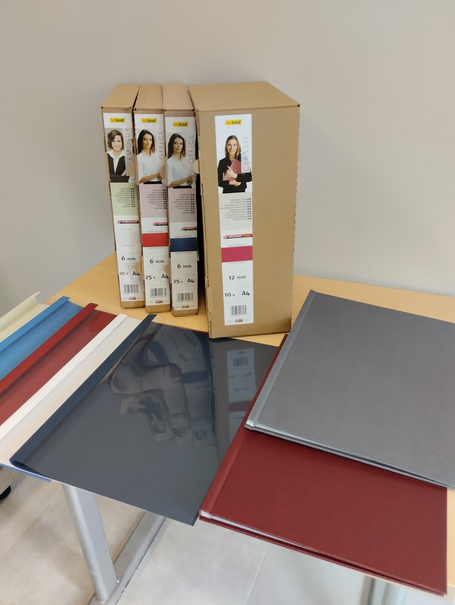 Pudełka z okładkami do bindowania Thermobind w różnych kolorach i grubościach, obok leżą pojedyncze okładki w kolorach: białym, czerwonym, niebieskim, szarym i granatowym.