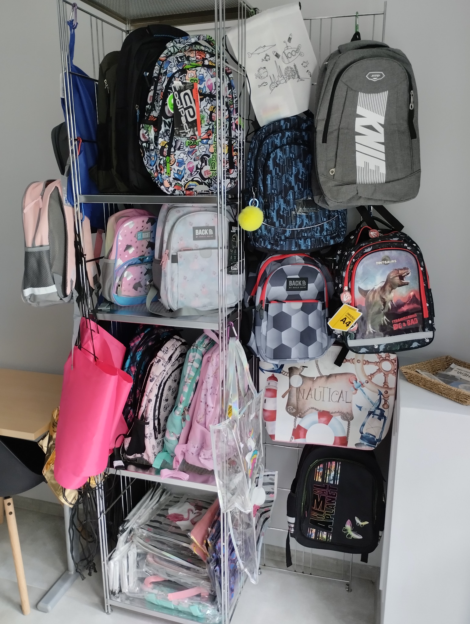 Ekspozycja różnorodnych plecaków szkolnych na metalowym regale, widoczne różne wzory i kolory, od plecaków z motywami zwierzęcymi po geometryczne wzory i napisy.