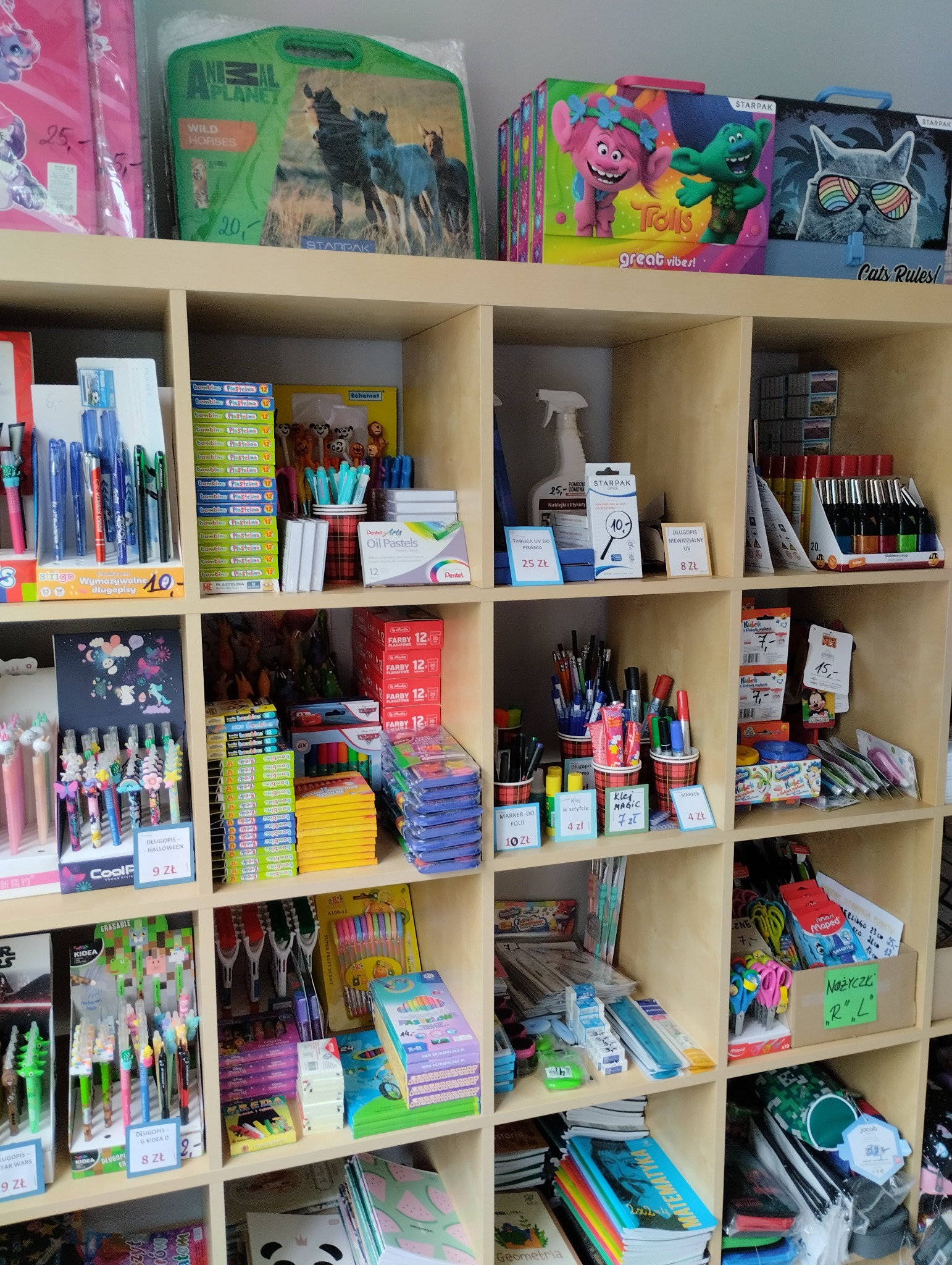 Półki sklepowe z artykułami papierniczymi i szkolnymi, w tym długopisy, kredki, farby, plastelina, zeszyty i akcesoria biurowe, ułożone tematycznie i cenowo.