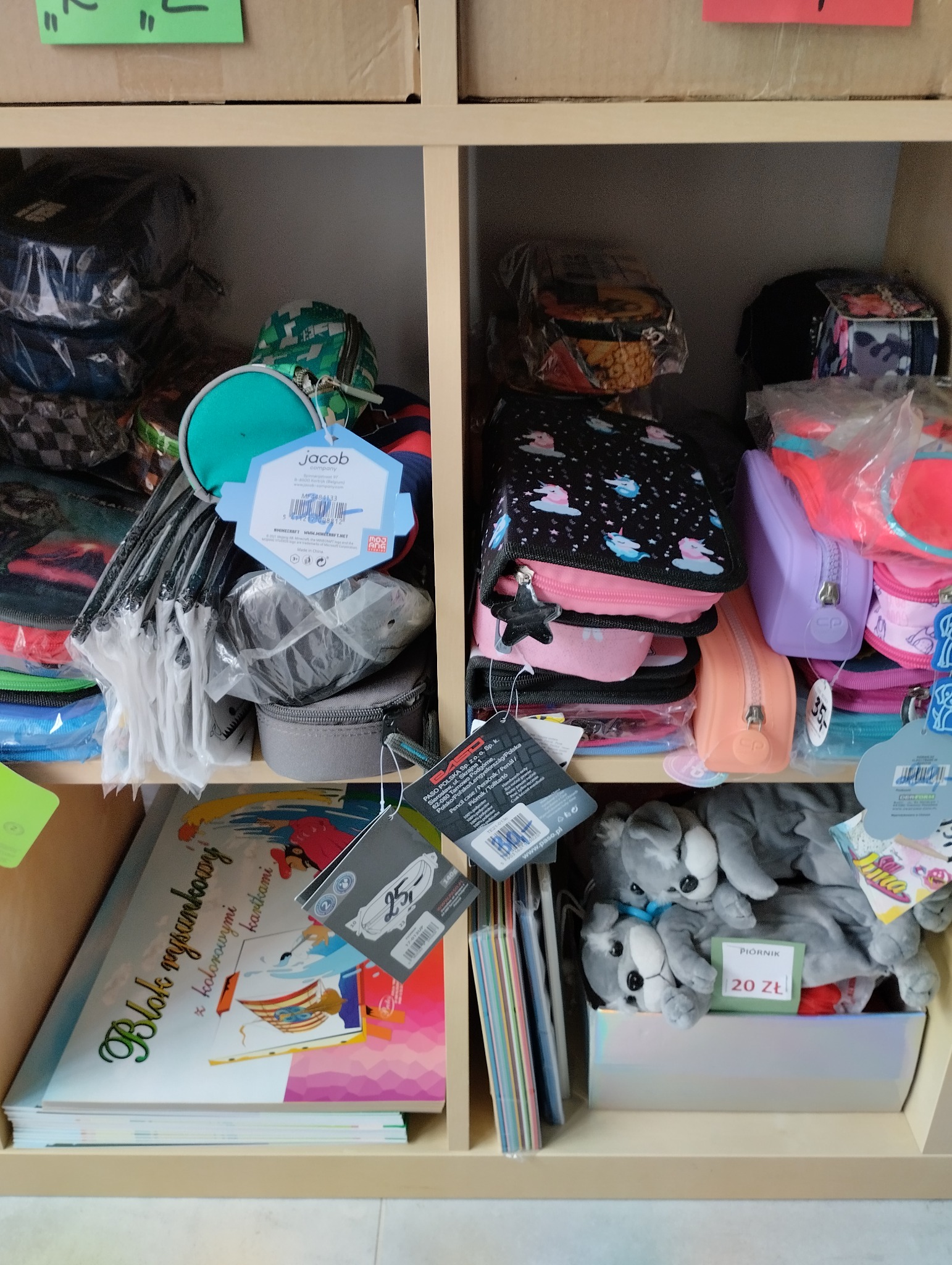 Półka z artykułami szkolnymi: piórniki z zawieszką w kształcie gwiazdki, bloki rysunkowe, kolorowe długopisy i pluszowe piórniki w kształcie piesków z ceną 20 zł.
