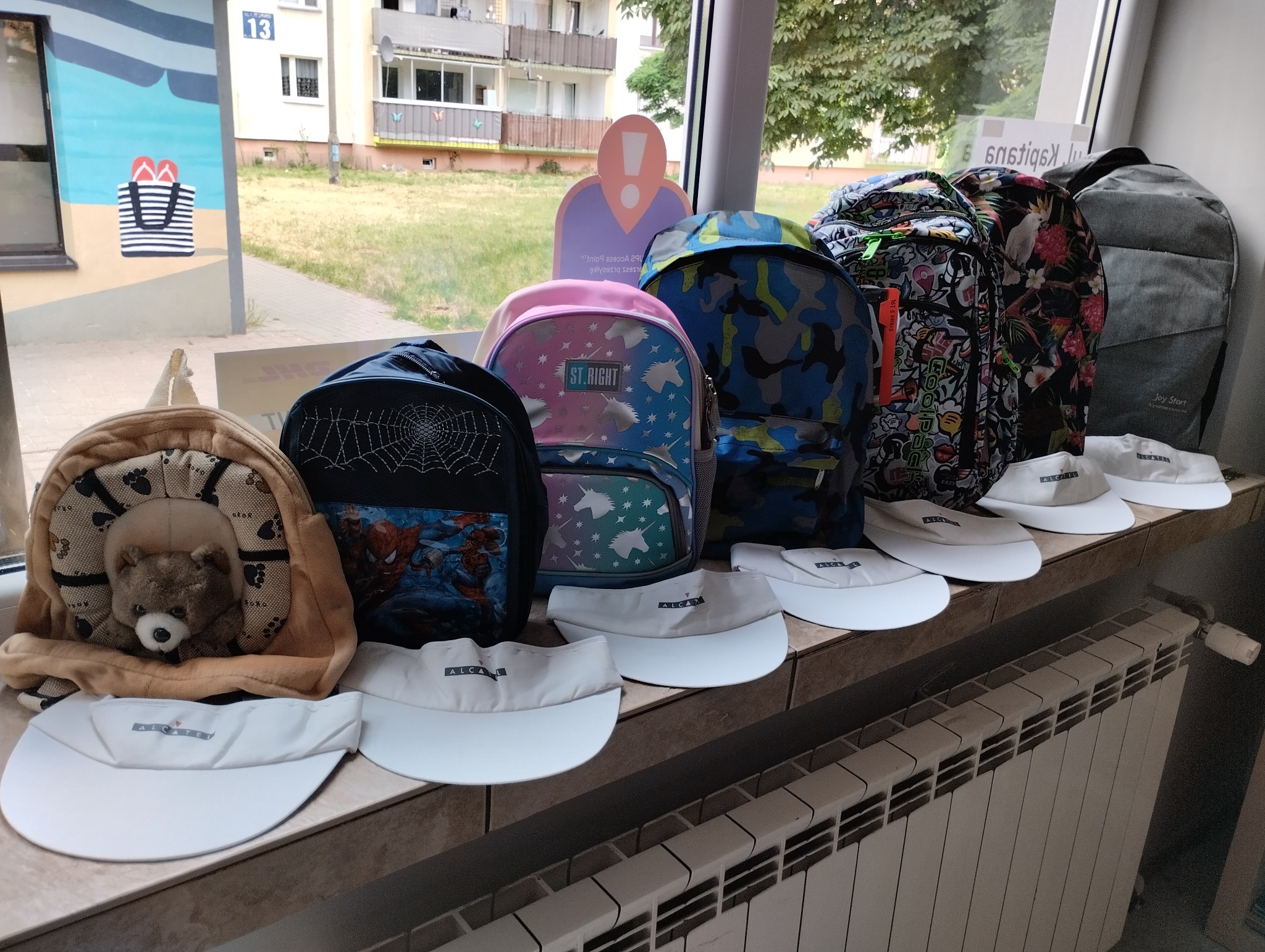Różnorodne plecaki dziecięce z motywami Spider-Mana, jednorożców i kamuflażu, ustawione obok siebie na parapecie okiennym, z rzędem białych daszków z logo 'ALCATEL' ułożonych przed nimi, w tle...