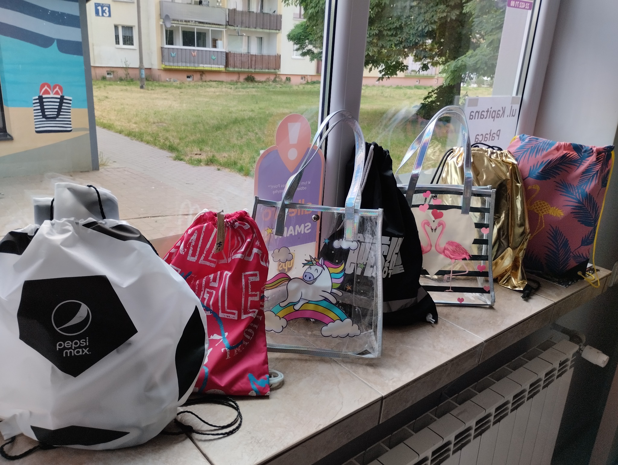 Różnokolorowe torby i worki z różnymi wzorami, w tym piłka nożna Pepsi Max, jednorożec na tęczy, flamingi i liście palmowe, ustawione na parapecie okiennym z widokiem na budynek mieszkalny.