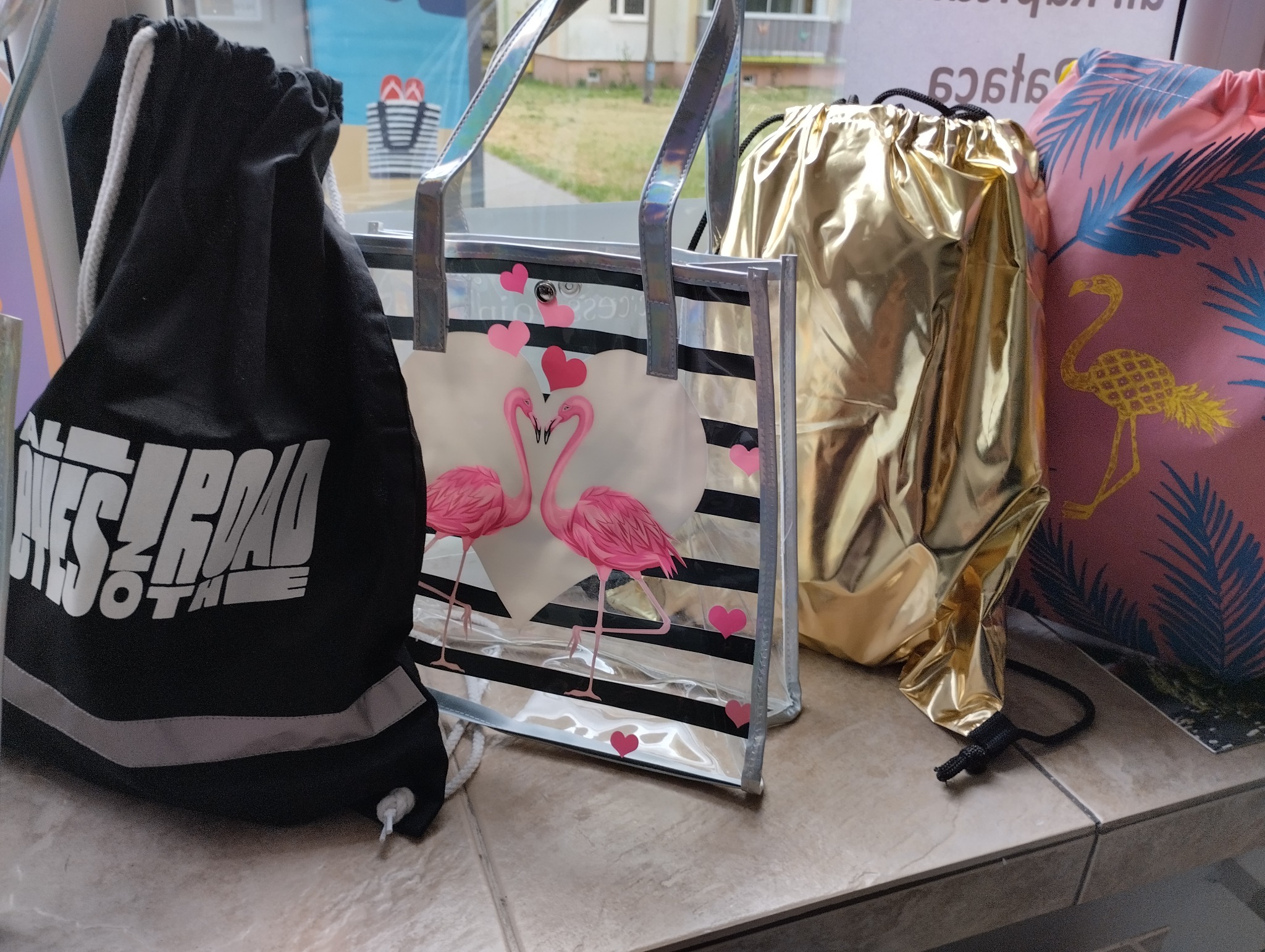 Cztery torby i worki: czarny worek gimnastyczny z napisem, przezroczysta torba na zakupy w paski z motywem flamingów i serc, złoty worek ze sznurkiem i różowy worek z motywem flaminga i liści...