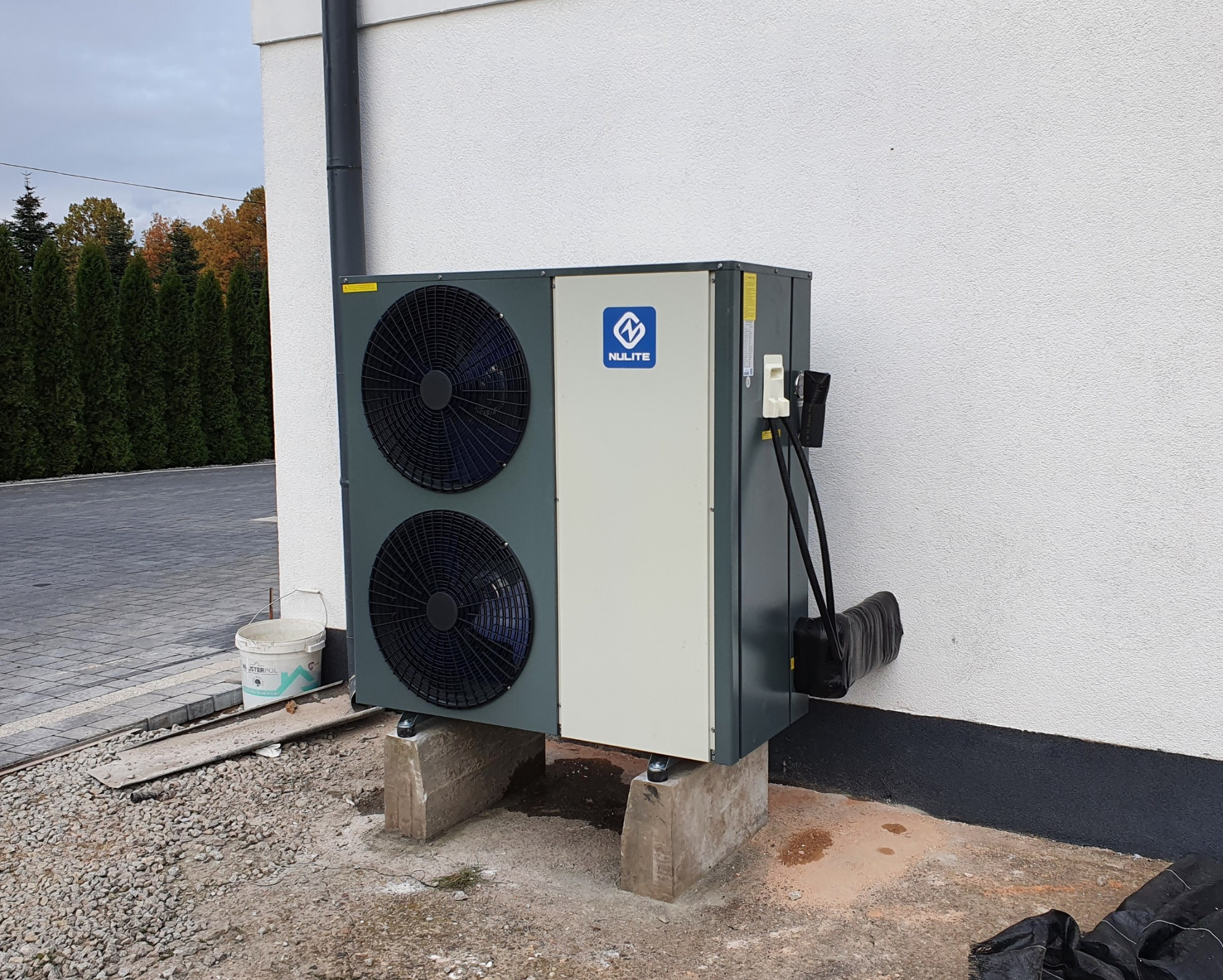 Zewnętrzna jednostka pompy ciepła marki NULITE, zamontowana na betonowych podstawach przy białej ścianie budynku, widok z boku, z widocznym przewodem odprowadzającym skropliny.