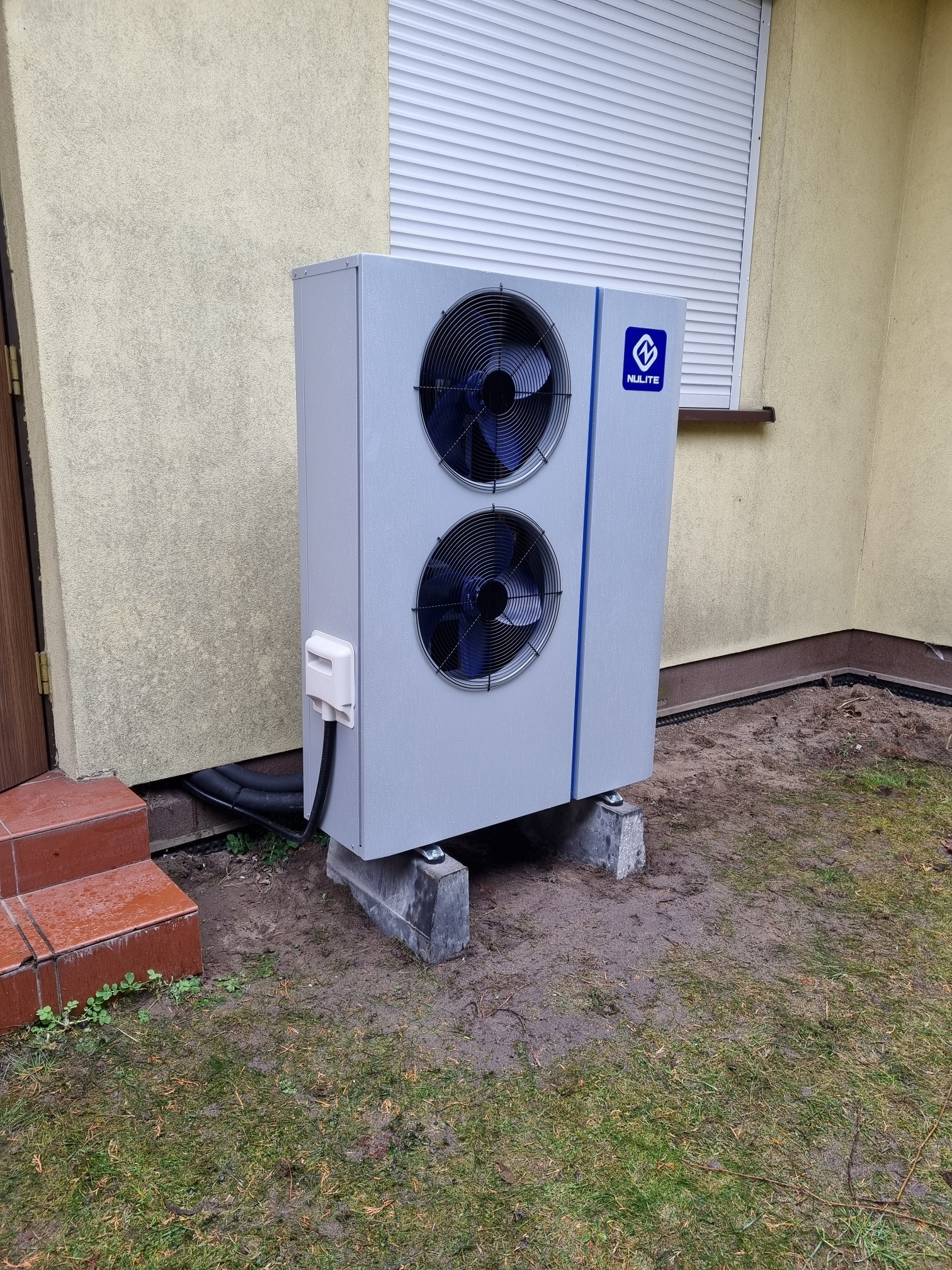 Zewnętrzna jednostka szarej pompy ciepła z dwoma wentylatorami, umieszczona na betonowych bloczkach przy ścianie budynku z roletami, widoczne przyłącza elektryczne i hydrauliczne.