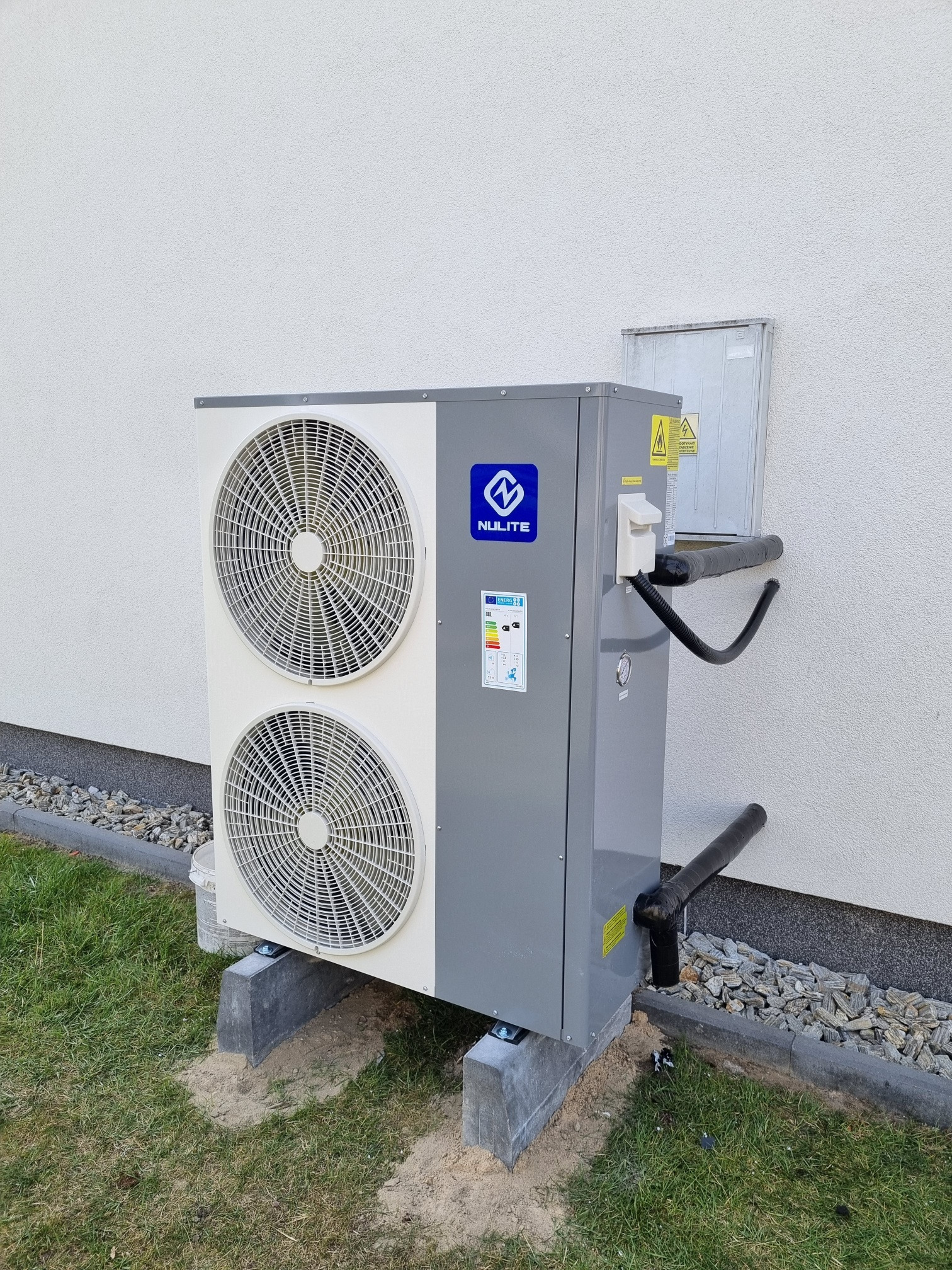 Zewnętrzna jednostka szaro-białej pompy ciepła marki Nulite, zamontowana na betonowych podstawach przy jasnej ścianie budynku, z widocznymi rurami odprowadzającymi skropliny i kostką brukową w tle.