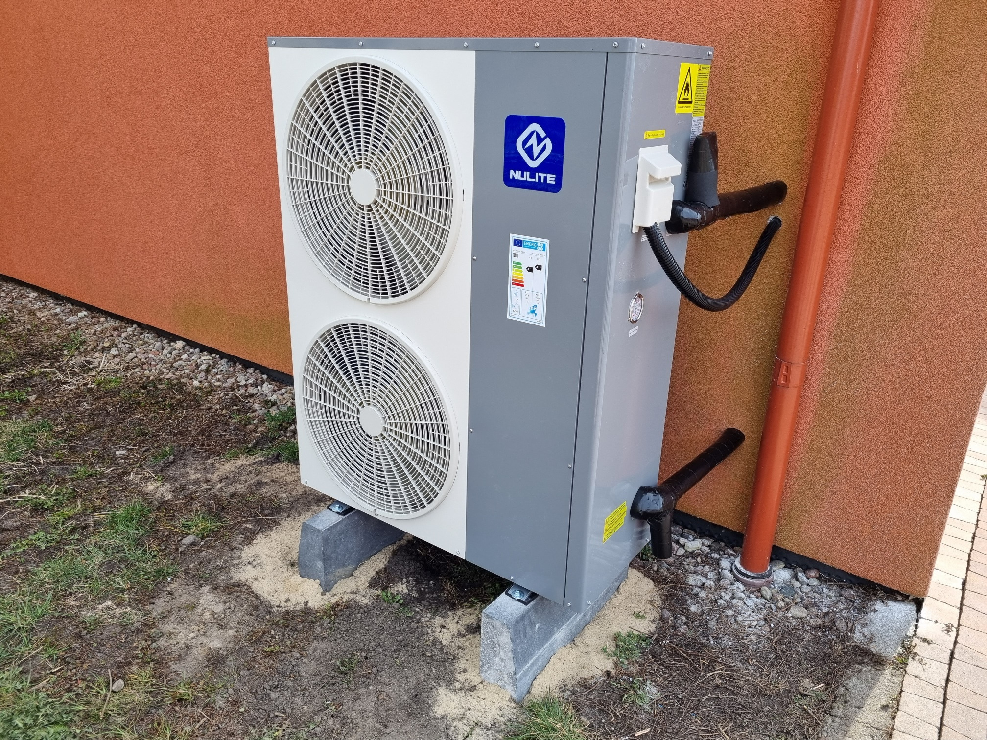 Zewnętrzna jednostka pompy ciepła Nulite, zamontowana na betonowych podstawach przy ścianie budynku o pomarańczowej elewacji, widoczna rura spustowa.