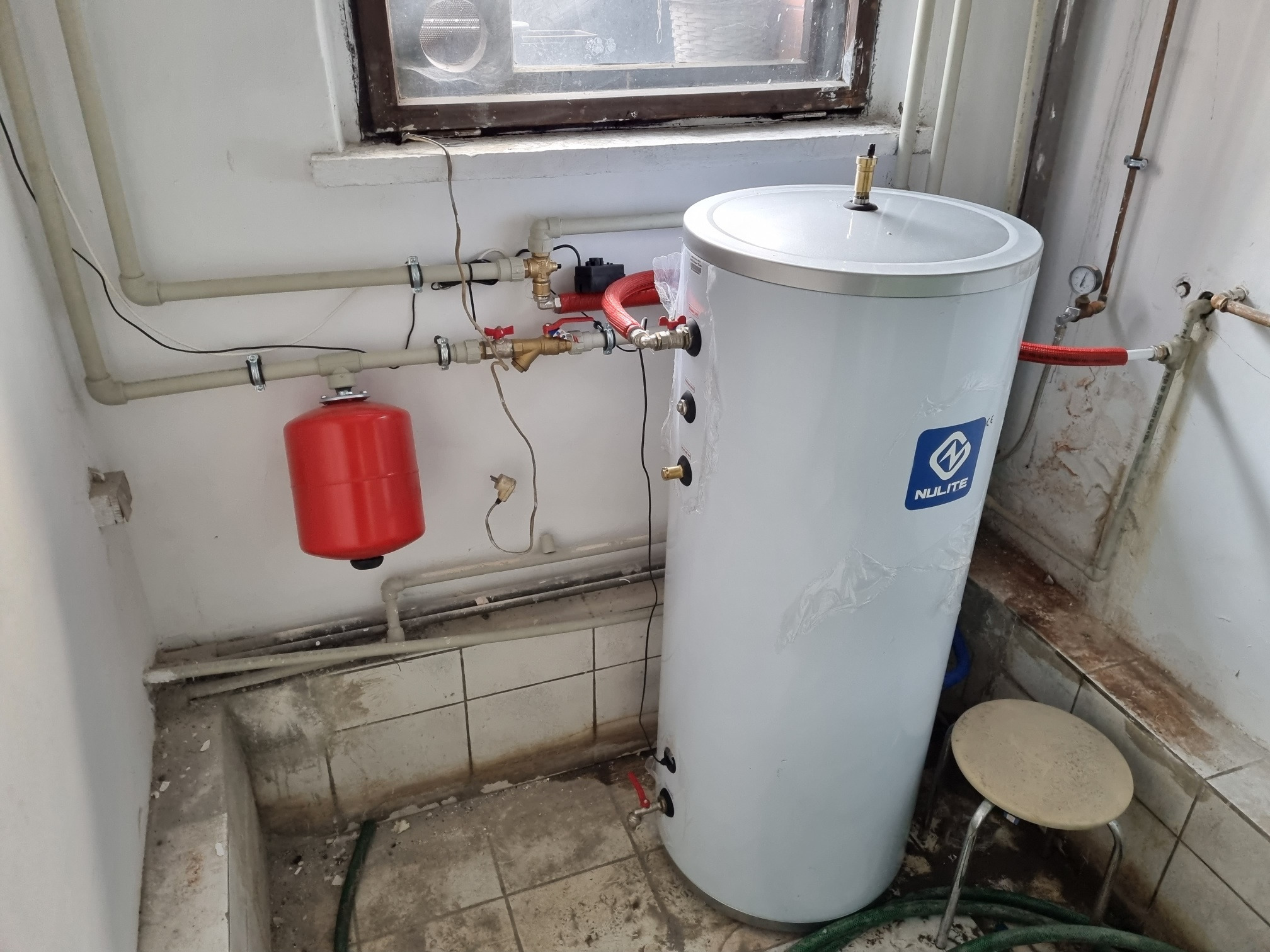 Bojler z podłączoną instalacją hydrauliczną w pomieszczeniu gospodarczym, widoczny czerwony zbiornik wyrównawczy i rury z zaworami, obok stoi mały taboret.