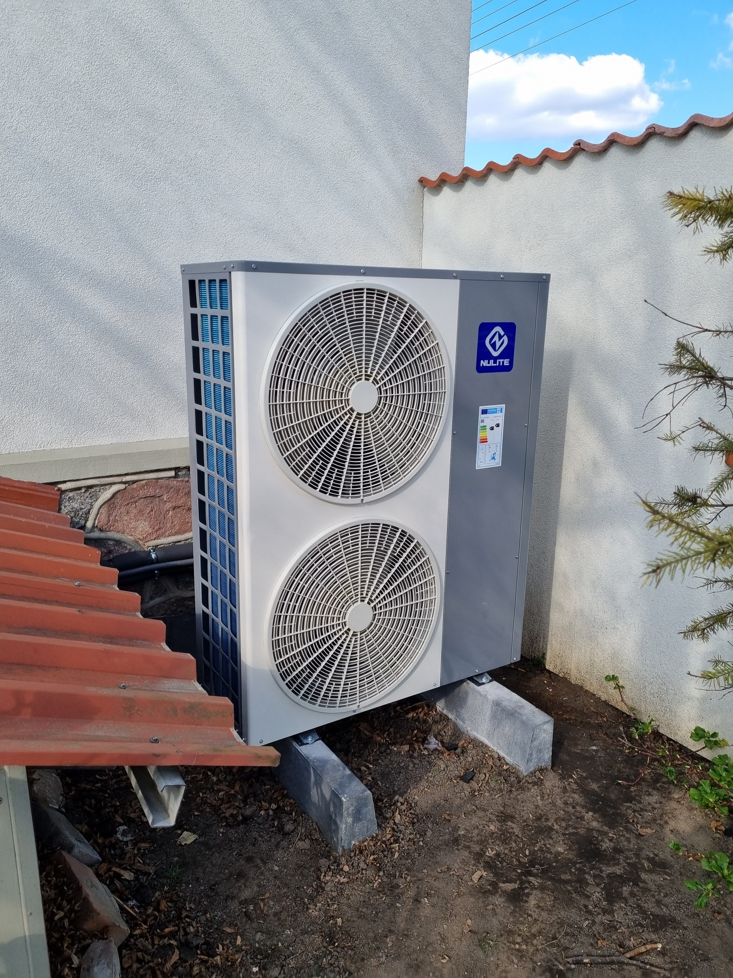 Szara jednostka zewnętrzna z dwoma wentylatorami marki Nulite, zamontowana na betonowych podstawach przy białej ścianie budynku z pomarańczową dachówką na krawędzi, widoczna instalacja i fragment...