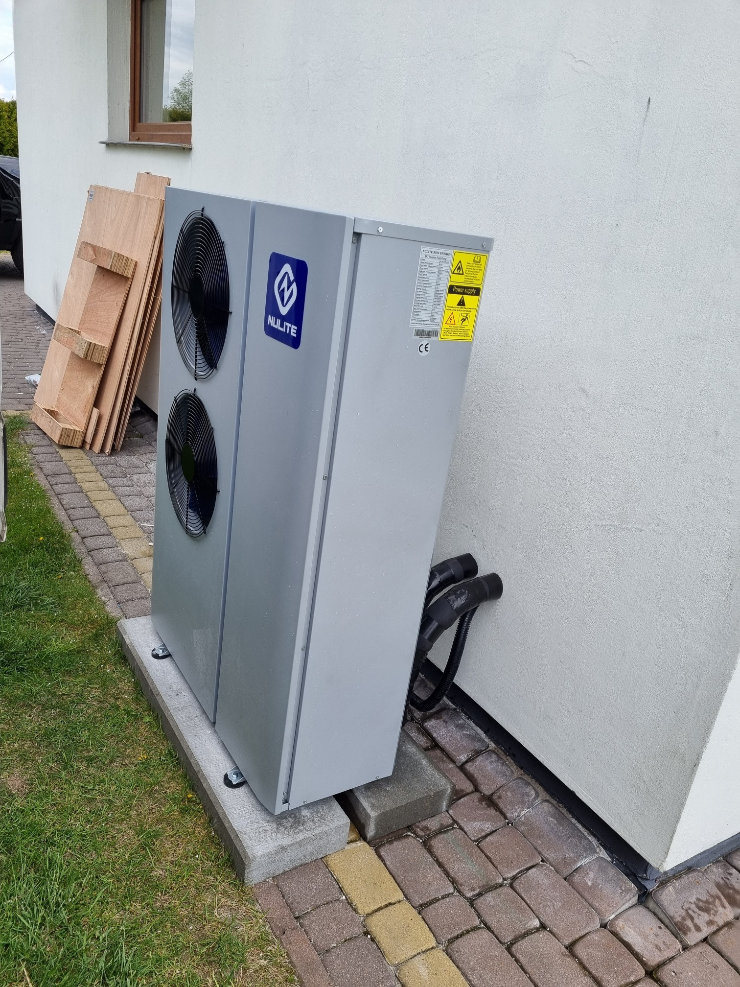 Szara pompa ciepła marki Nulite zainstalowana na betonowej podstawie przy białej ścianie budynku, widoczne przewody i paleta drewniana w tle.