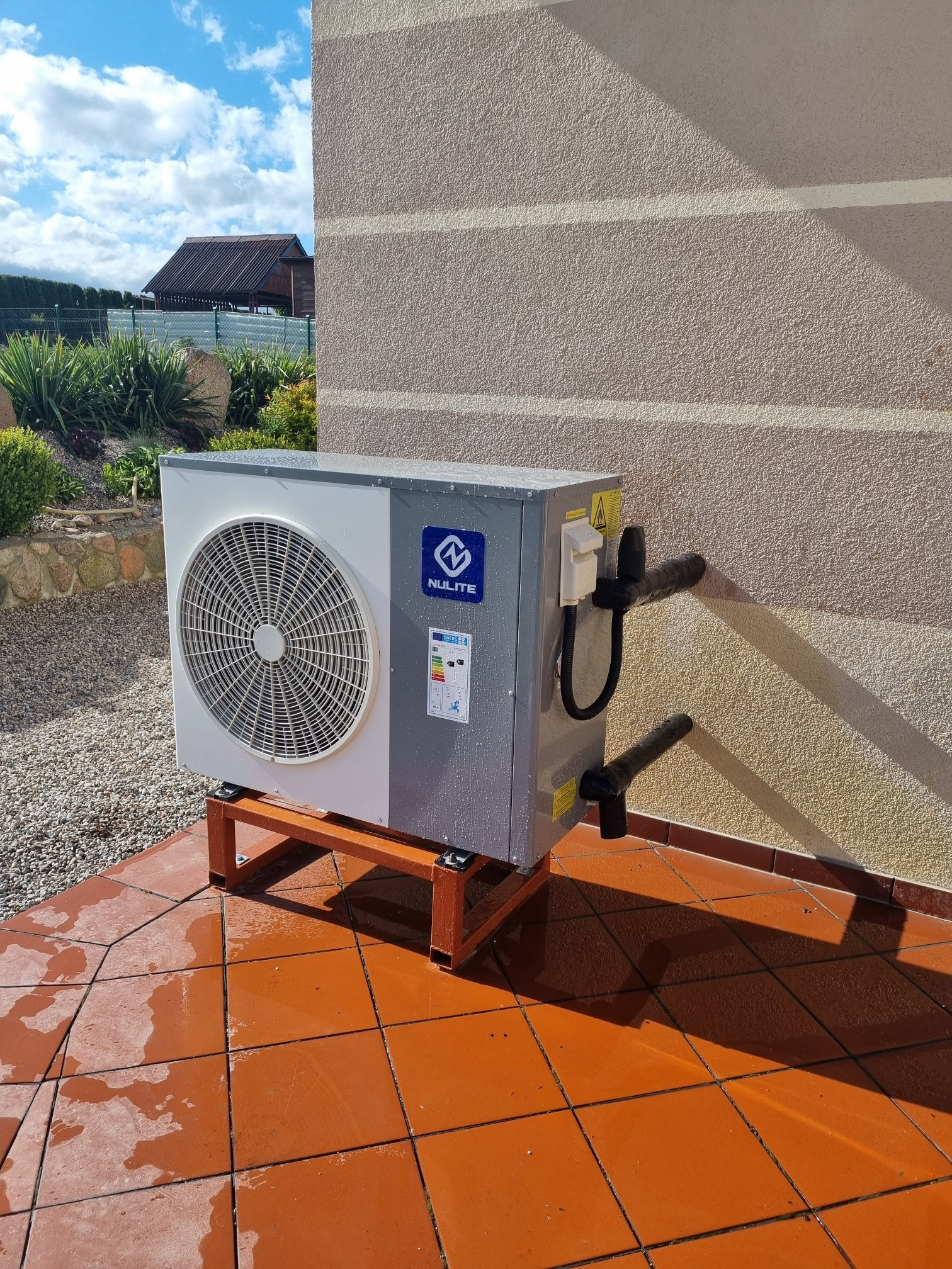 Zewnętrzna jednostka szaro-białej pompy ciepła marki Nulite, umieszczona na pomarańczowym, metalowym stojaku, przy ścianie budynku z elewacją w kolorze beżowym, widoczne rury i przewody, lekko...