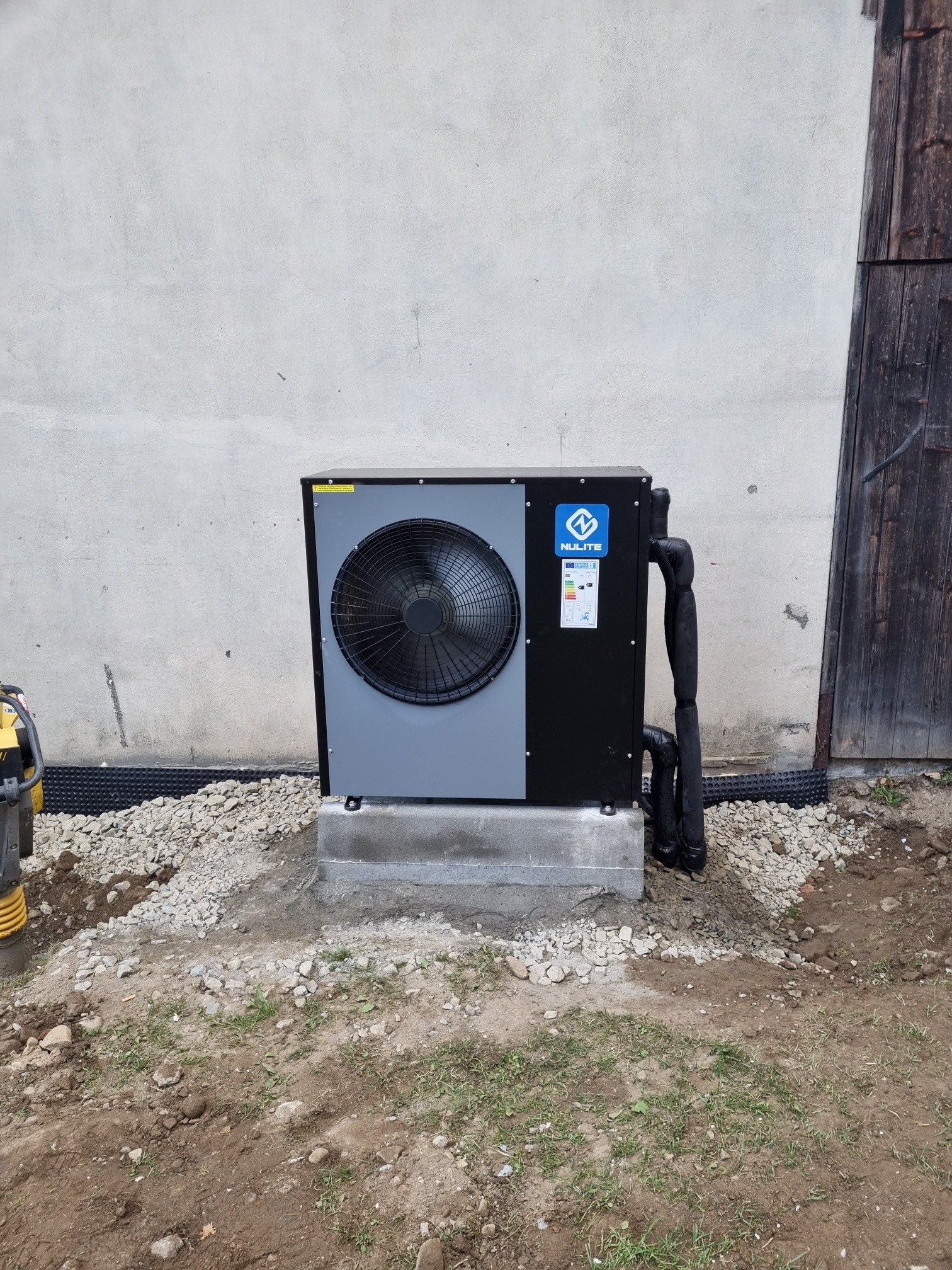 Szaro-czarna jednostka zewnętrzna z wentylatorem, marki Nulite, zainstalowana na betonowej podstawie, z czarnymi rurami, na tle białej ściany i drewnianych drzwi, z widoczną ubijarką w tle.