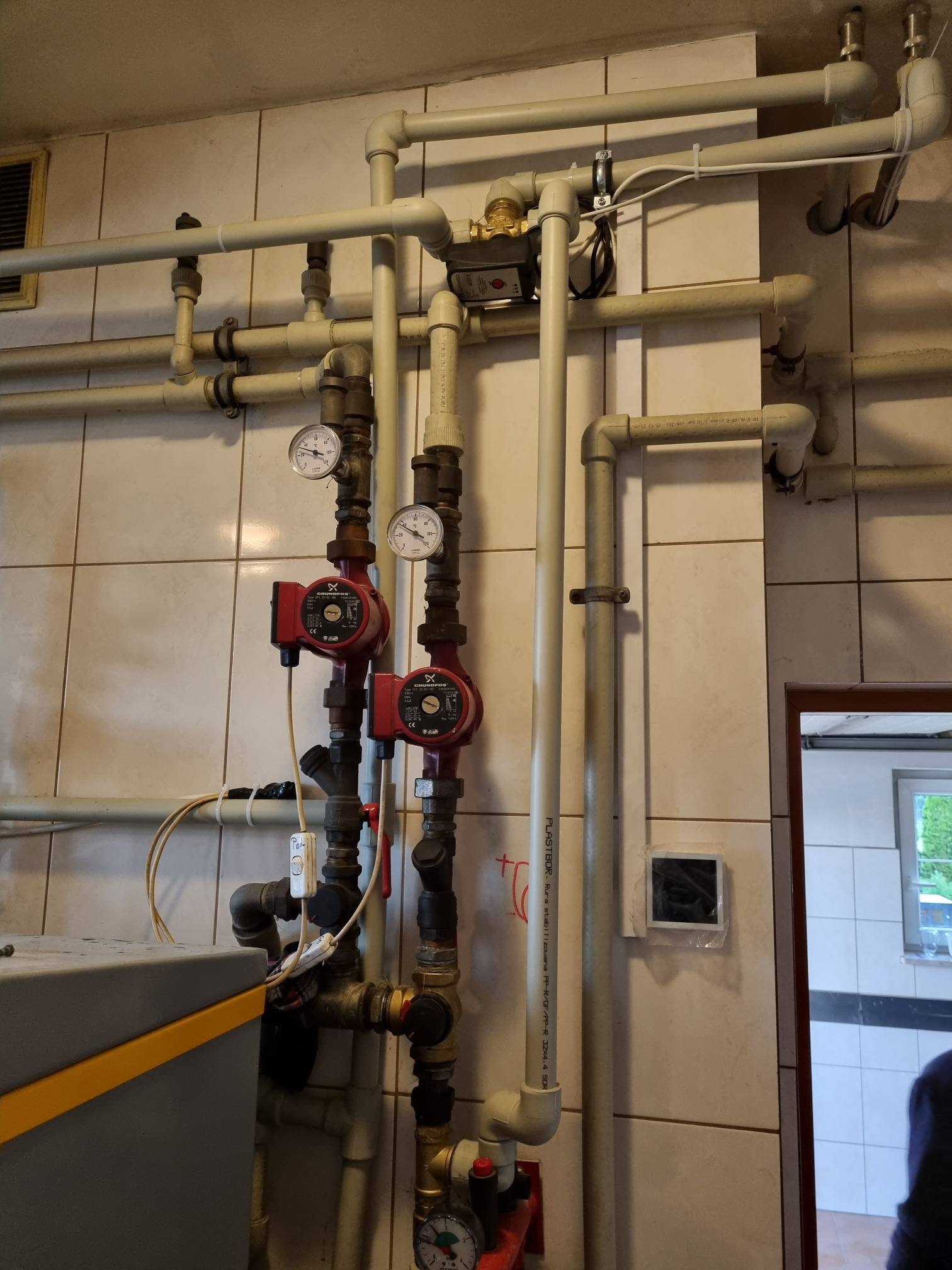 Instalacja rur z dwoma czerwonymi pompami obiegowymi marki Grundfos z manometrami, zamontowana na ścianie z płytek ceramicznych, widoczne liczne złączki i zawory.