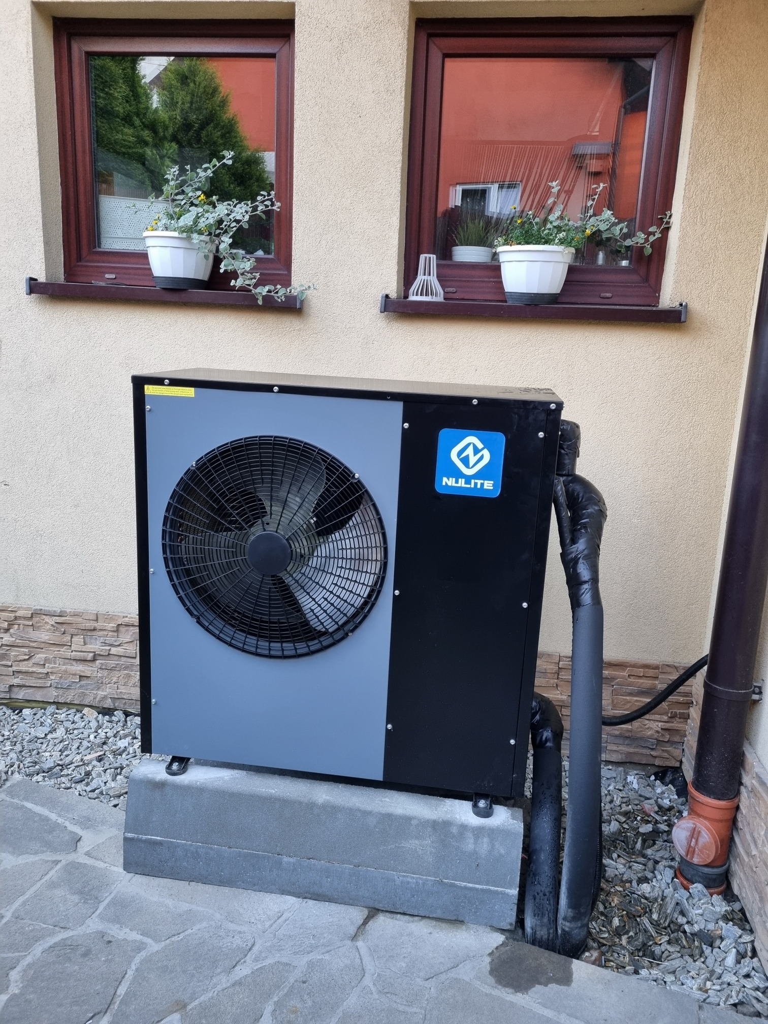 Zewnętrzna jednostka pompy ciepła marki Nulite, umieszczona na betonowym fundamencie przed budynkiem z dwoma oknami, na których parapetach stoją doniczki z roślinami.