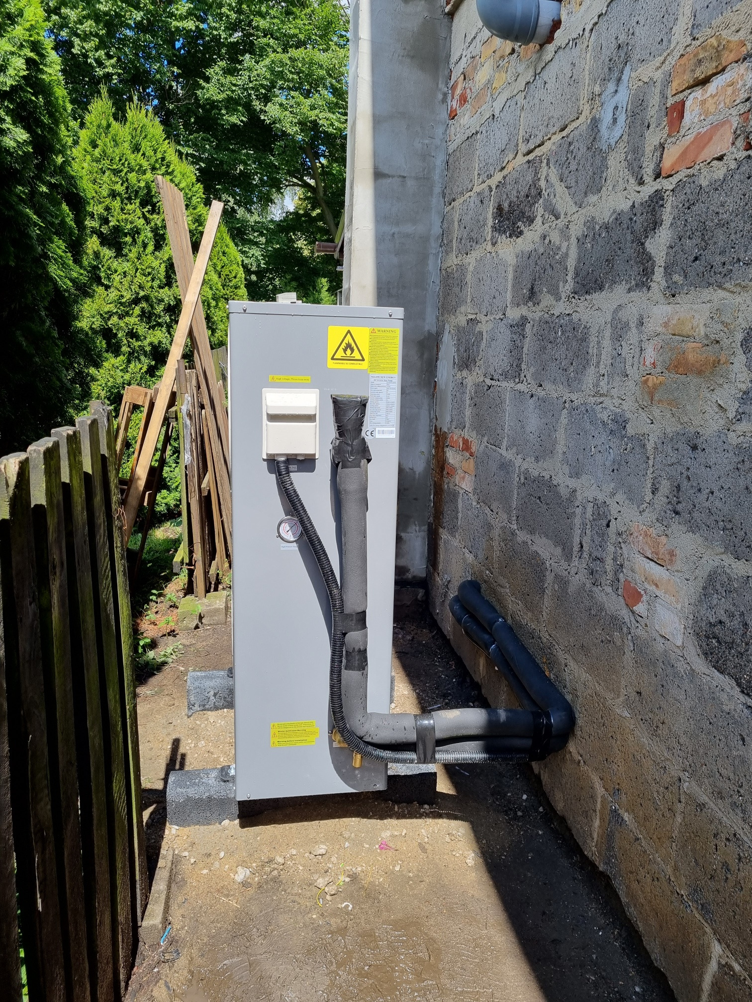 Szara pompa ciepła zamontowana przy ceglanym budynku w Łomiankach, widoczne czarne rury przyłączeniowe i tabliczka ostrzegawcza o wysokim napięciu.