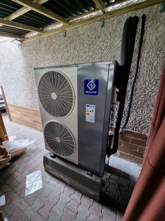 Zewnętrzna jednostka pompy ciepła Nulite z dwoma wentylatorami, zamontowana na betonowej podstawie przy ścianie z szorstkim tynkiem pod zadaszeniem.