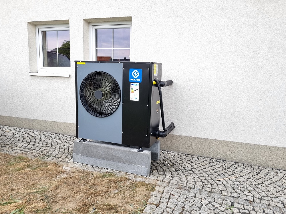 Zewnętrzna jednostka pompy ciepła NULITE, zamontowana na betonowych blokach przy ścianie budynku z dwoma oknami, widoczne rury przyłączeniowe i kostka brukowa wokół.