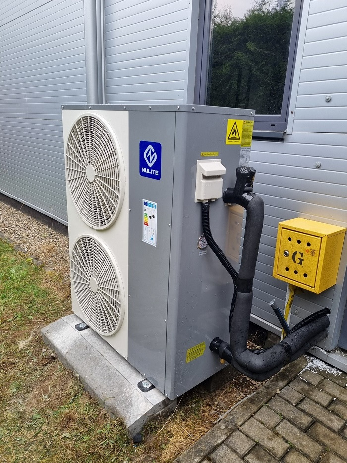 Zewnętrzna jednostka klimatyzacyjna z dwiema turbinami, zamontowana na betonowym fundamencie obok ściany budynku z żółtą skrzynką gazową, widoczny fragment okna i trawnika.