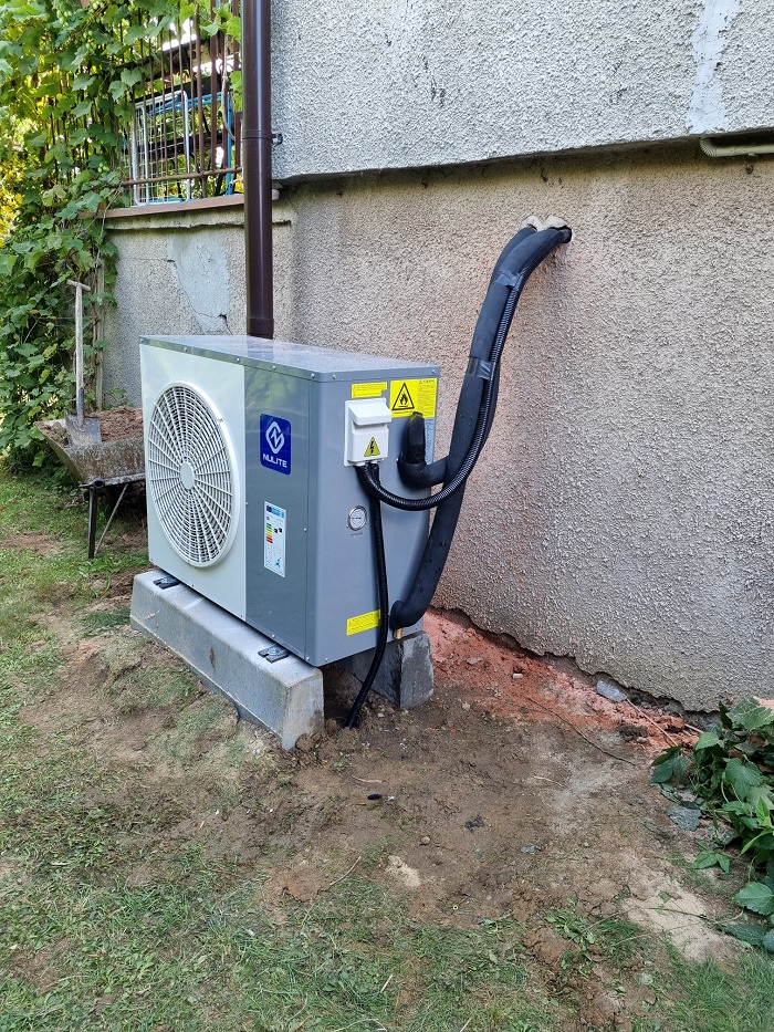 Szara pompa ciepła marki NULITE zainstalowana na betonowych bloczkach przy ścianie budynku z widocznymi rurami w izolacji, w tle zielona roślinność i częściowo widoczny wózek z łopatą.