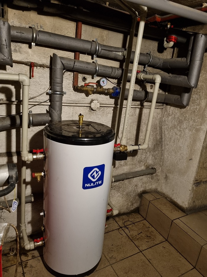 Biały zbiornik buforowy Nulite w kotłowni z widocznymi rurami i zaworami na tle szarej ściany.