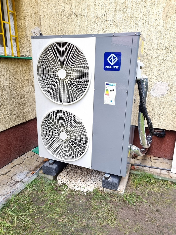 Zewnętrzna jednostka pompy ciepła Nulite, zainstalowana na betonowych bloczkach i podsypana białym grysem przy ścianie budynku w Łomiankach, z widocznymi przewodami w izolacji.