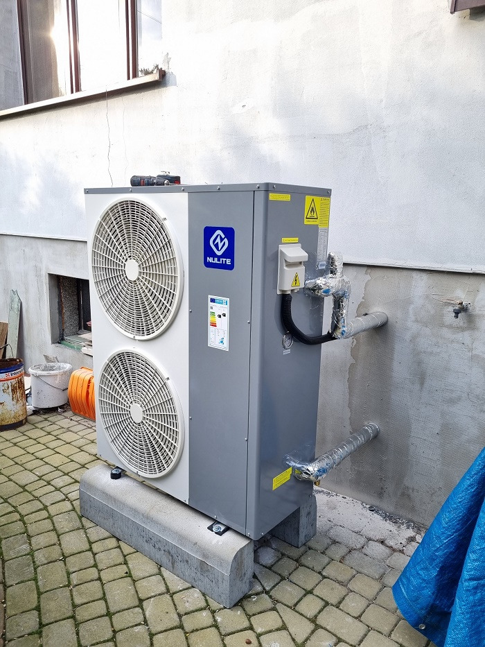 Zewnętrzna jednostka szarej pompy ciepła Nulite, zamontowana na betonowych podstawach, z widocznymi rurami w izolacji, na tle jasnej ściany budynku i kostki brukowej.
