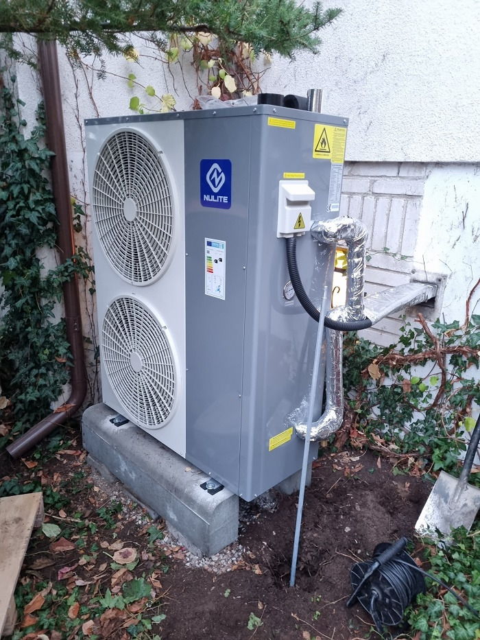 Zewnętrzna jednostka pompy ciepła Nulite, zamontowana na betonowej podstawie przy ścianie budynku z widocznymi rurami w izolacji aluminiowej i kablem zasilającym na ziemi.