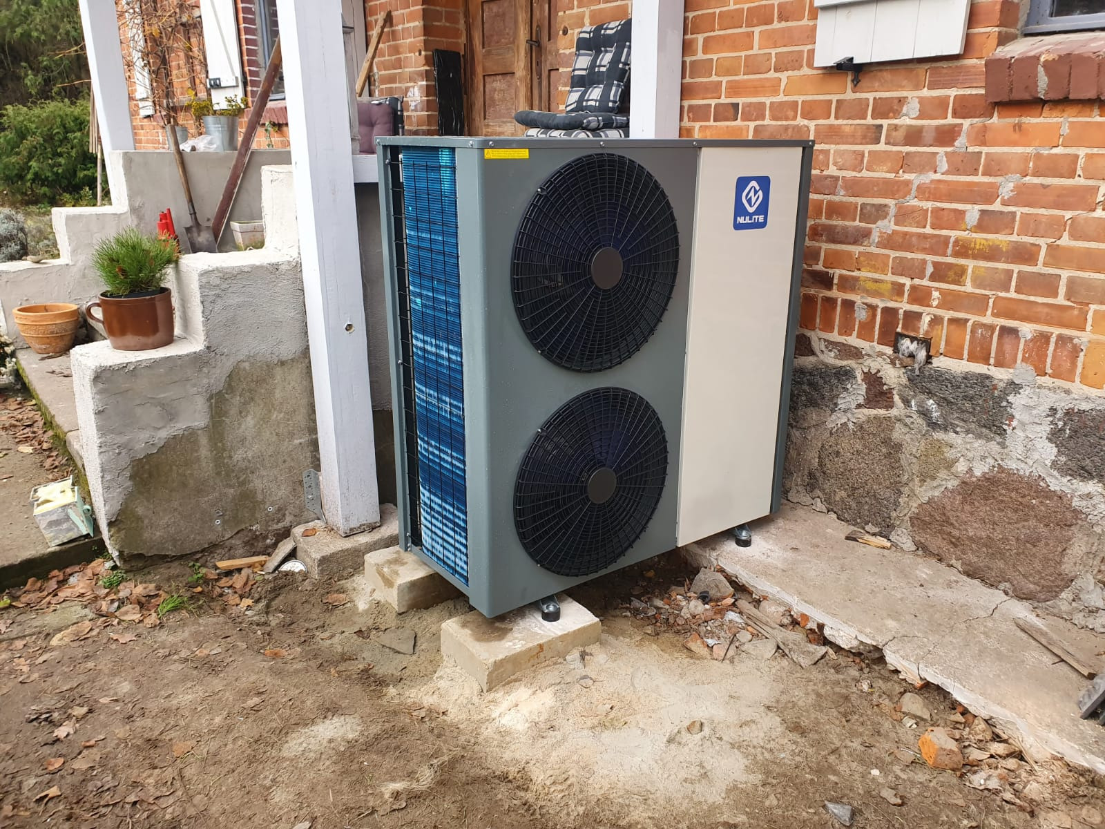 Zewnętrzna jednostka z dwoma wentylatorami zamontowana na betonowych blokach przy ceglanym budynku, widoczny fragment schodów z doniczkami.