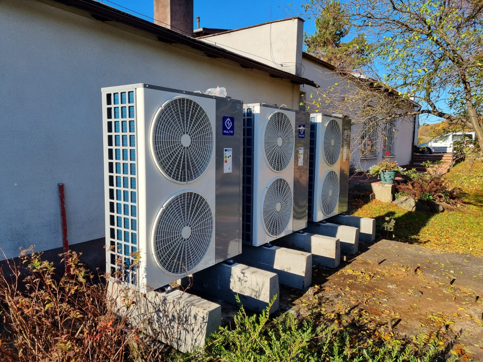 Cztery jednostki zewnętrzne, prawdopodobnie agregaty chłodnicze, ustawione rzędem na betonowych podstawach przy ścianie budynku w słoneczny dzień. Widoczna marka NULITE na każdym urządzeniu.