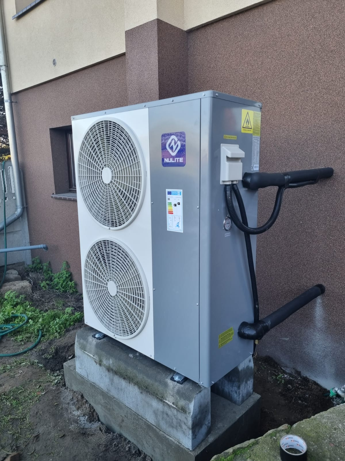 Zewnętrzna jednostka z dwoma wentylatorami marki Nulite, zamontowana na betonowym fundamencie przy ścianie budynku z brązowym tynkiem strukturalnym, widoczne rury izolowane termicznie.