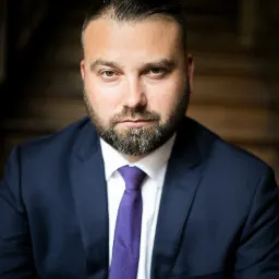 Portret mężczyzny z brodą w ciemnym garniturze i fioletowym krawacie, patrzącego w obiektyw na tle drewnianych schodów.