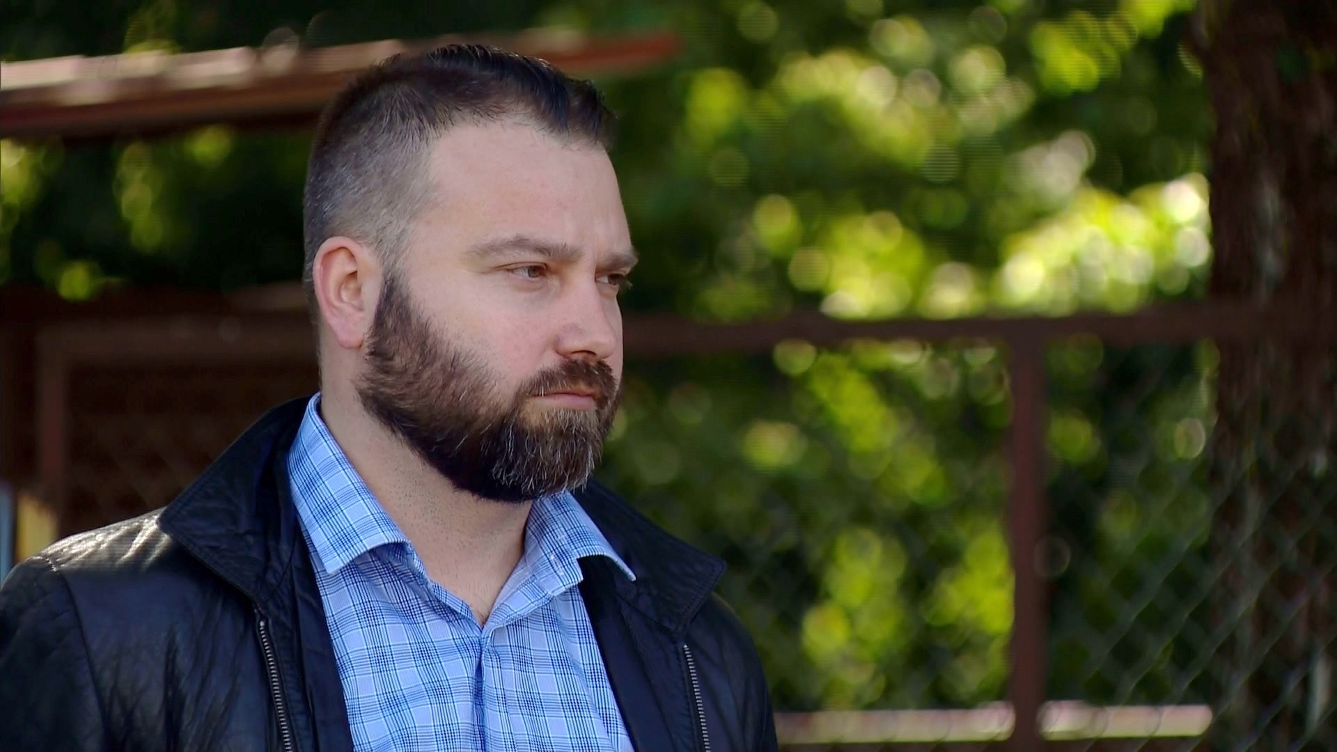 Portret zamyślonego mężczyzny z brodą, w niebieskiej koszuli w kratę i czarnej skórzanej kurtce, spoglądającego w lewo, na tle rozmytej zieleni.