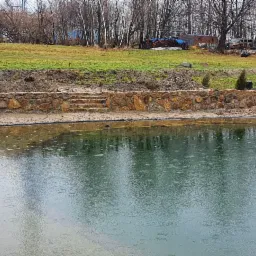 Oczko wodne z kamiennym brzegiem i schodami z kamienia, w tle trawnik i budynek z drewna.
