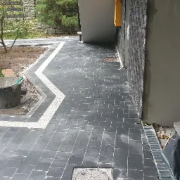 Ułożona, ciemnoszara kostka brukowa z jasną obwódką wzdłuż budynku i ogrodu, widoczny kubeł z narzędziami i odpływ liniowy.