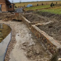 Budowa kamiennego murku przy stawie w ogrodzie, widoczne wykopane rowy, błoto i drewniany dom w tle.