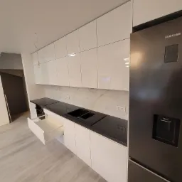 Nowoczesna kuchnia z białymi, połyskującymi frontami szafek i czarnym blatem, uzupełniona o lodówkę Samsung w ciemnym kolorze. Otwarta szuflada ukazuje białe wnętrze. Na ścianie białe płytki...