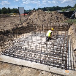 SBK-Instal - Pracownik w żółtym kasku i kamizelce układa zbrojenie w wykopie fundamentowym, widoczne stalowe pręty tworzące szkielet konstrukcji, w tle hałda ziemi i błękitne niebo z chmurami.