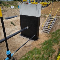 SBK-Instal - Betonowa konstrukcja z izolacją bitumiczną, rurami i instalacją gazową w wykopie, metalowe schody z żółtymi poręczami prowadzą w dół, widoczne taśmy ostrzegawcze.