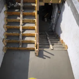 Wylana betonowa podłoga z drewnianym szalunkiem schodów, widok z góry na klatkę schodową w surowym stanie, białe ściany z bloczków betonowych, etap budowy.