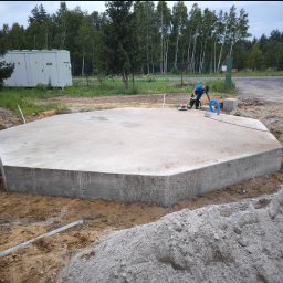 SBK-Instal - Wieloboczna płyta betonowa, prawdopodobnie pod budowę, otoczona wykopem i stertami ziemi, z pracownikiem używającym narzędzia do obróbki powierzchni, transformator w tle.