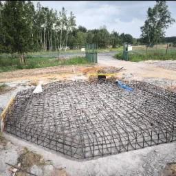 Szkielet z prętów zbrojeniowych w wykopie, przygotowany do zalania betonem, z widocznymi deskami szalunkowymi i poziomnicą w tle, na tle zieleni i pochmurnego nieba.