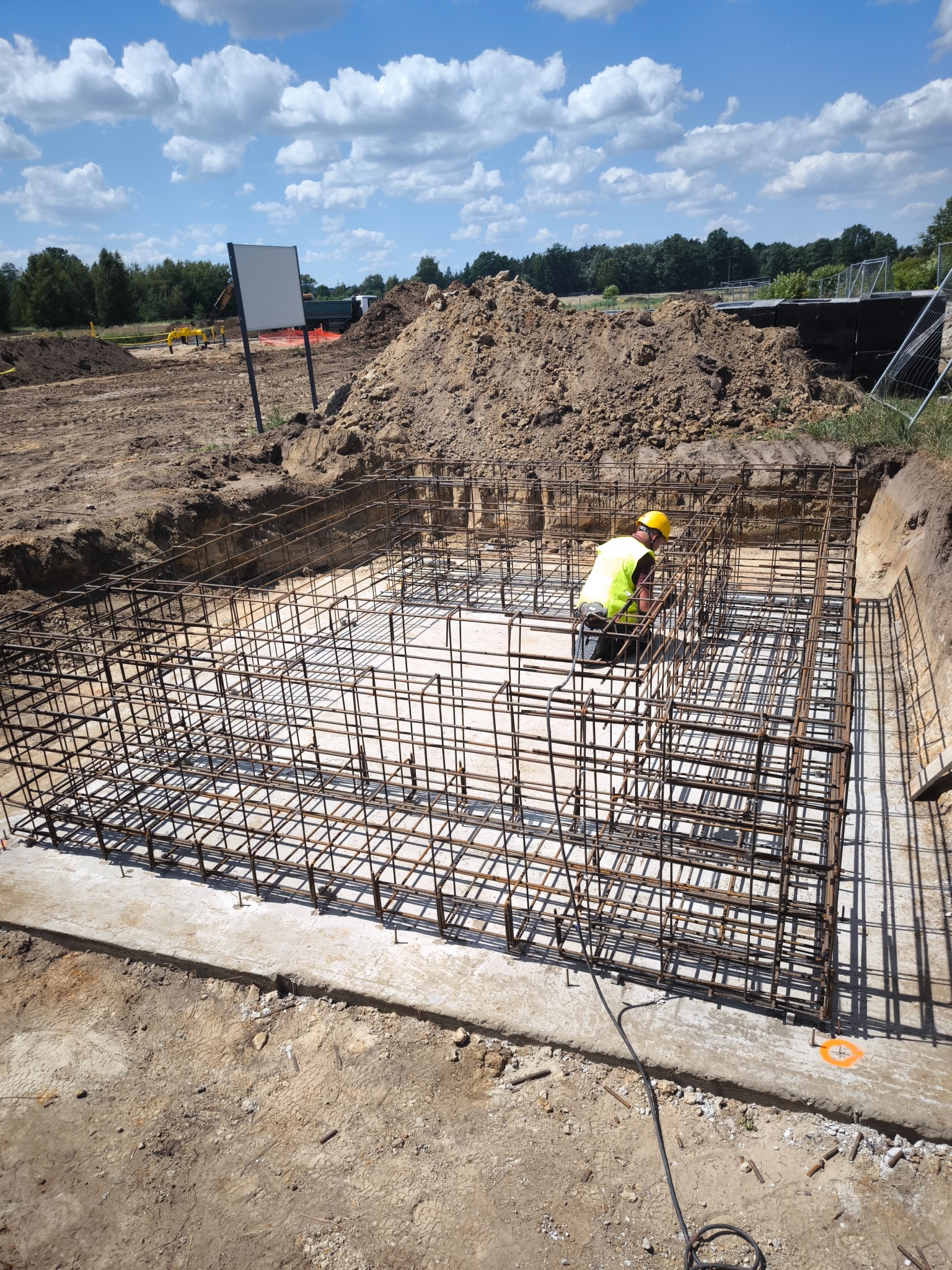 Pracownik w żółtym kasku i kamizelce układa zbrojenie w wykopie fundamentowym, widoczne stalowe pręty tworzące szkielet konstrukcji, w tle hałda ziemi i błękitne niebo z chmurami.